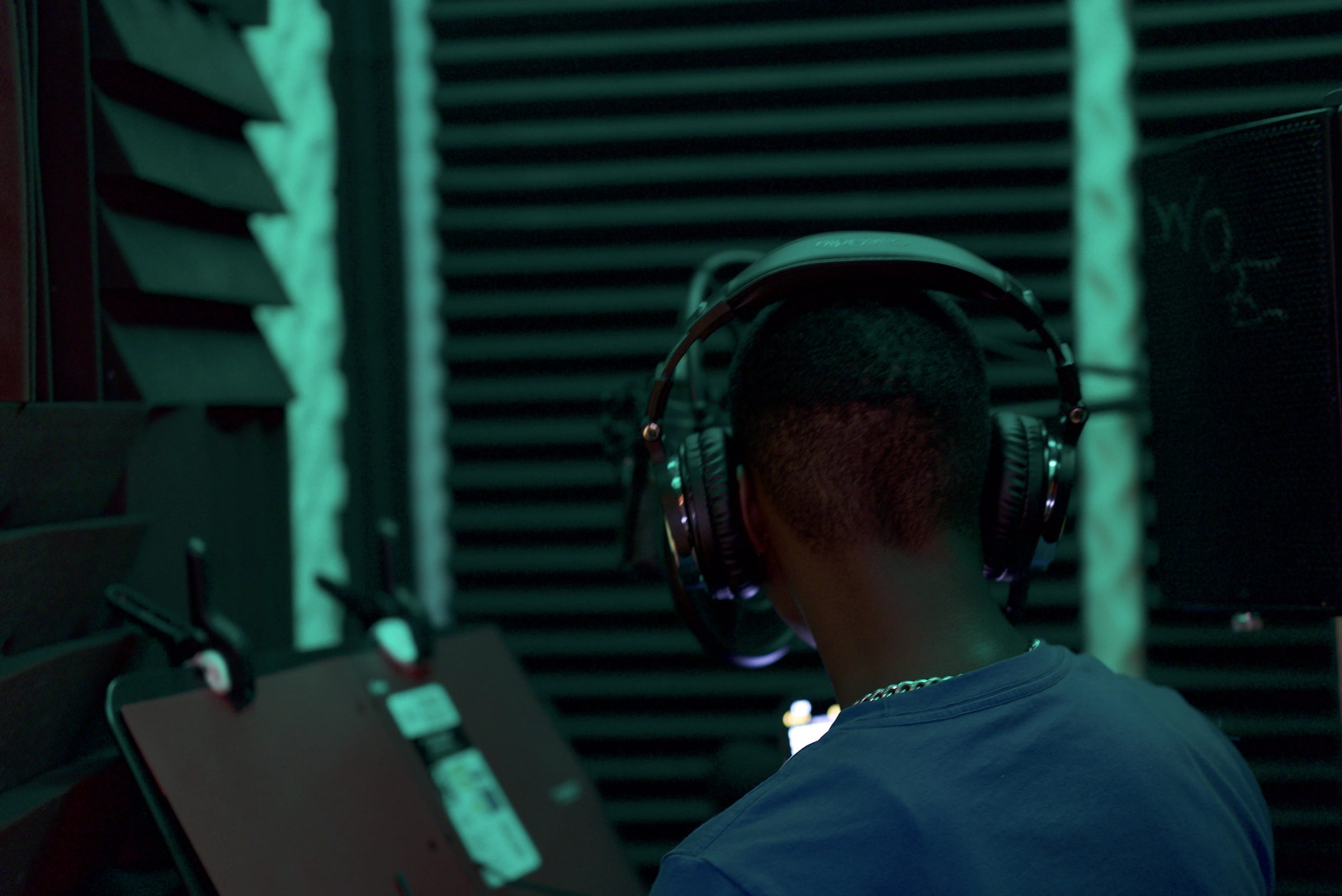 Artist in headphones recording in the Studio B vocal booth under blue-green light, seen from behind